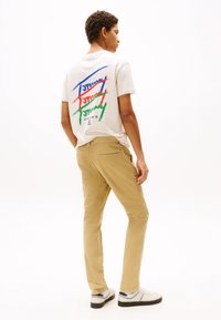 Camiseta blanca con texto colorido en la parte trasera y pantalones beige. El modelo lleva zapatillas blancas con detalles azules. Tejido suave, diseño casual.