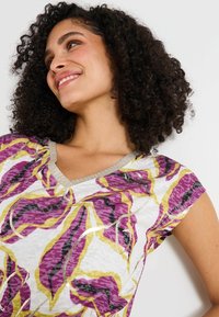 Top de manga corta y cuello en V en blanco con patrones de hojas amarillas, moradas y negras. Detalle en plata a lo largo del escote. Tejido texturizado.