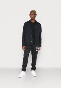 Sort jakke med to frontlommer, sort t-shirt med logo, sorte sweatpants med hvid "Nike" branding og hvide sneakers. Lyserøde solbriller.