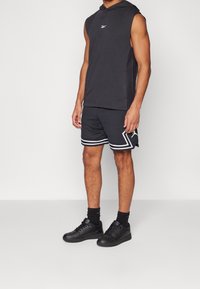 Hombre vestido con sudadera sin mangas negra, pantalones cortos de baloncesto negros con rayas blancas, calcetines negros y zapatillas negras, de pie sobre un fondo liso.