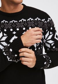 Black knit sweater featuring white snowflake pattern on chest and sleeves, ribbed cuffs, and crew neckline. Soft texture, casual design.