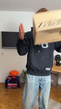 Kartonbox wird vorne gehalten, hellbraun mit bedrucktem schwarzen Text. Person trägt einen dunkelgrauen Kapuzenpullover und hellblaue Jeans in einem Raum mit Möbeln.