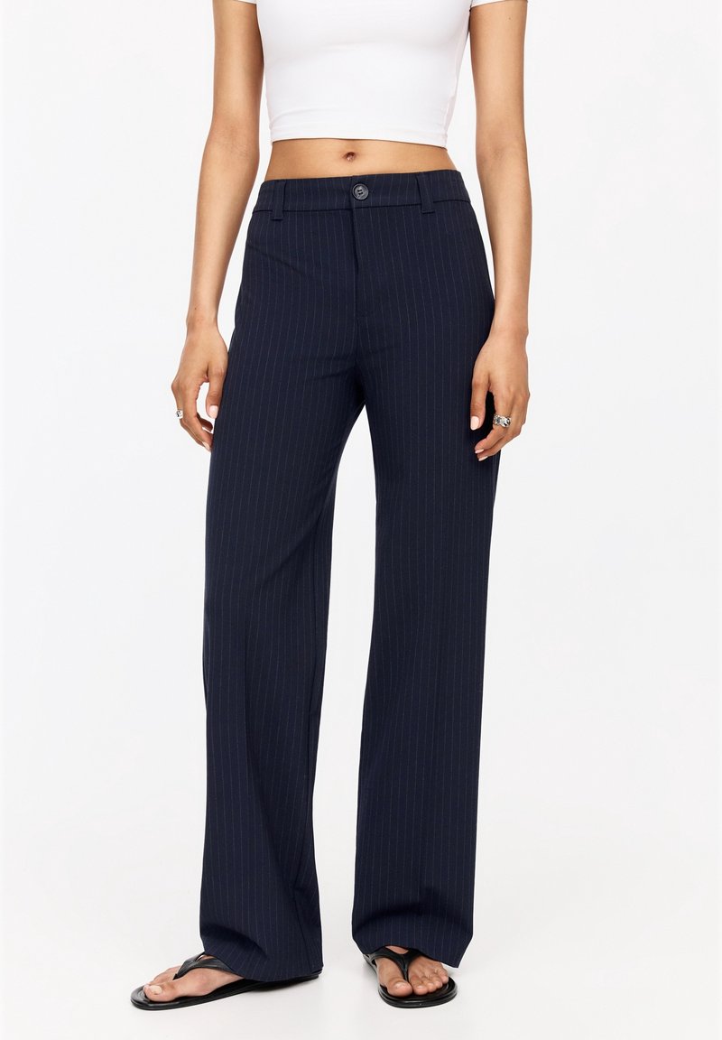 Femme portant un pantalon large à rayures noires, un top court blanc, des bagues aux doigts et des tongs noires, debout devant un fond blanc.