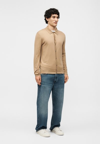 Νέος άντρας που στέκεται, φοράει ένα μπεζ πουκάμισο με κουμπιά, μπλε τζιν και λευκά αθλητικά παπούτσια σε απλό ανοιχτό φόντο.