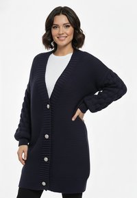 Granatowy dzianinowy kardigan z głębokim dekoltem w serek, teksturowanymi rękawami z wzorem warkoczowym oraz zapięciem na przodzie na zaokrąglone guziki.
