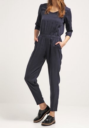 Femme portant une combinaison bleu marine à manches trois-quarts et des chaussures à lacets noires brillantes, les mains dans les poches, debout devant un fond uni.