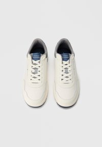 Λευκά αθλητικά παπούτσια Tommy Hilfiger με γκρι επένδυση, ναυτικό μπλε λεπτομέρειες και λευκά κορδόνια, εμφανιζόμενα από πάνω σε απλό φόντο.