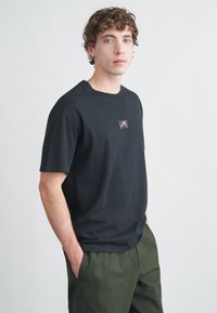 T-shirt preta de algodão com mangas curtas, com um pequeno patch gráfico no peito, combinado com calças verde-oliva. Textura suave.