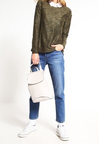 Oliivinvihreä teksturoitu villapaita, siniset suorat farkut, valkoiset lenkkarit ja vaaleanharmaa reppu, jossa on läppä ja pyöreät kahvat.