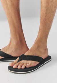 Tongs noires avec des brides texturées, dotées d'une semelle en caoutchouc et de lignes accentuées blanches. Le design est ouvert à l'avant pour une meilleure respirabilité.