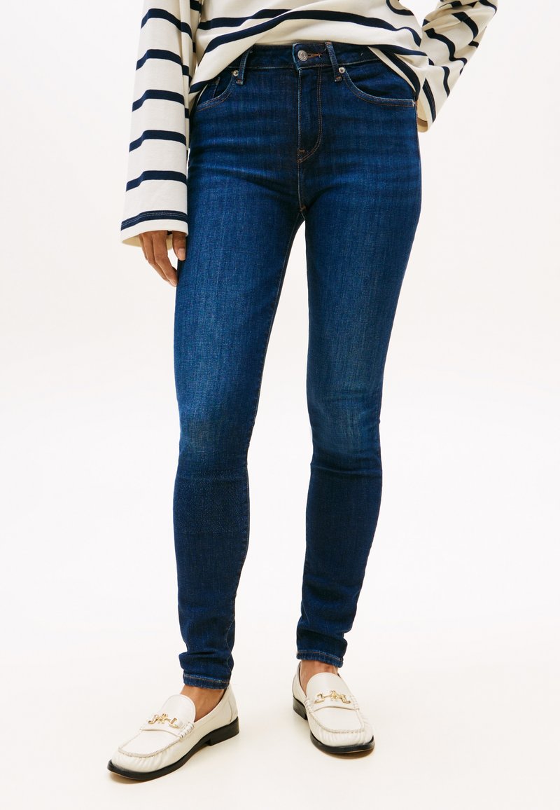 Person trägt dunkelblaue Skinny-Jeans, weiße Loafers mit goldfarbenen Details und ein weißes Langarmshirt mit schwarzen horizontalen Streifen.