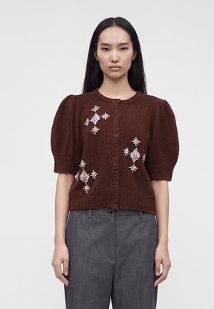 Kvinde iført en brun kortærmet cardigan med blomsterudsmykninger og grå højtaljede bukser foran en ensfarvet baggrund.