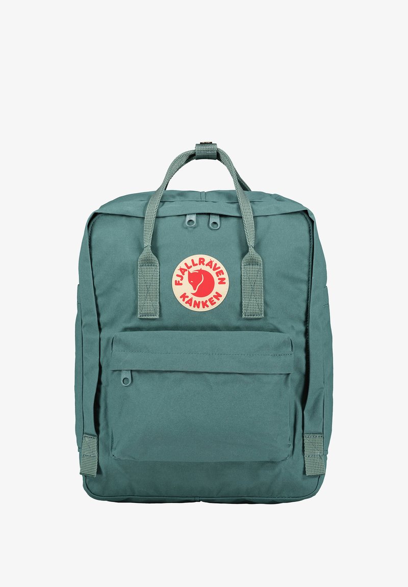 Mochila de tecido verde com formato retangular, etiqueta de logo, pega superior e alças ajustáveis. Possui um bolso frontal com fecho e material texturizado.
