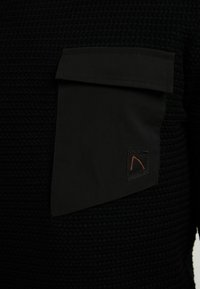 Černá texturovaná košile s velkou černou kapsou a jemným oranžovým logem v rohu. Materiál vypadá tkaný a robustní.