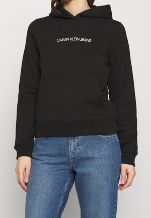 Czarna bluza z kapturem Calvin Klein Jeans z przednią kieszenią, noszona przez osobę w niebieskich dżinsach, przycięta na wysokości szyi i bioder.