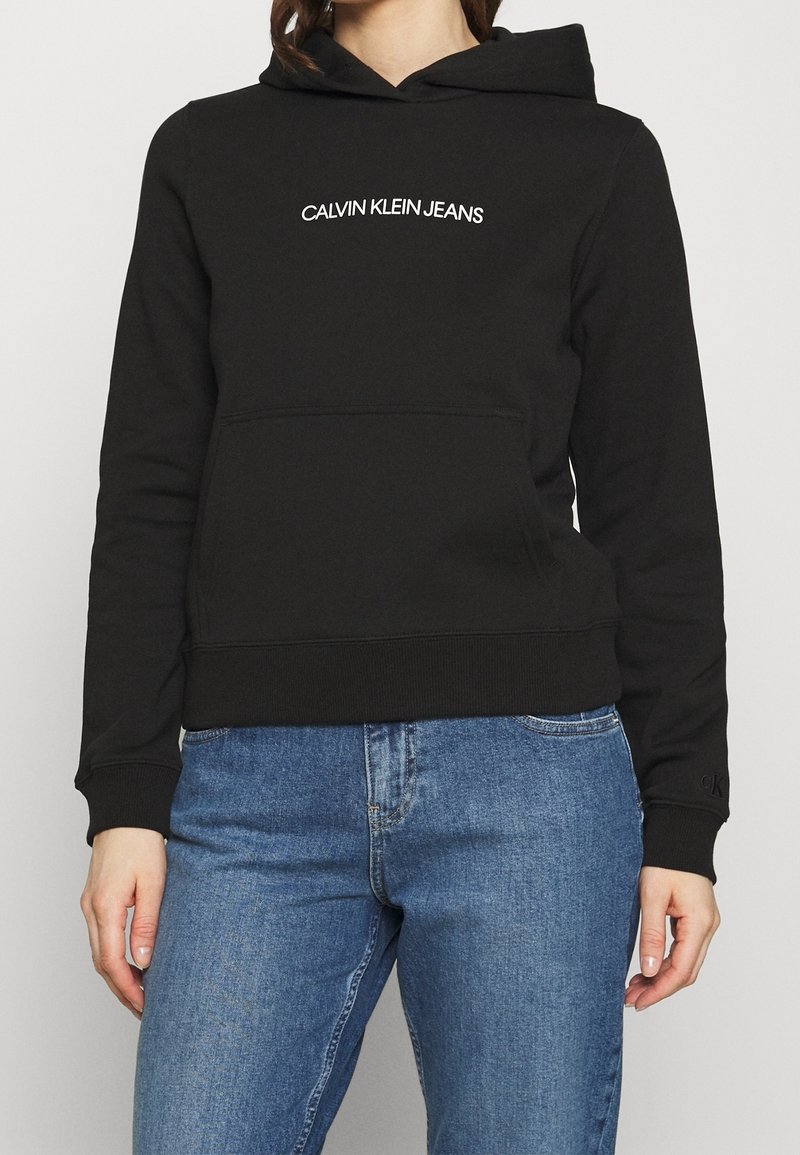 Sweat à capuche noir Calvin Klein Jeans avec poche avant, porté par une personne en jean bleu, recadré au niveau du cou et des hanches.