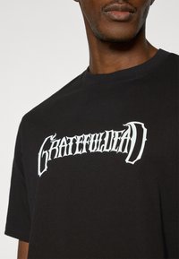 Μαύρο βαμβακερό μπλουζάκι με έντονα λευκά γράμματα "GRATEFUL" σε γοτθική γραμματοσειρά. Στρογγυλή λαιμόκοψη και κοντά μανίκια. Ομαλή υφή.