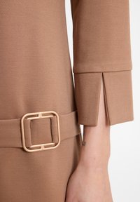 Nahaufnahme eines beigen Mantelärmel mit einem Schlitzbund und einem passenden Stoffgürtel mit einer quadratischen Schnalle in der Taille.