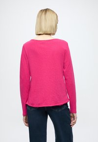 Roza majica z dolgimi rokavi in sproščenim krojem. Mehka tekstura, izrez z okroglim vratom in ukrivljen rob. Opravljena z vidnimi temno modrimi kavbojkami na pasu.