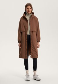 Bruine, lichte, gewatteerde jas met een relaxte pasvorm, elastische manchetten en een ritsluiting, gedragen over een crèmekleurige gebreide trui en zwarte leggings.