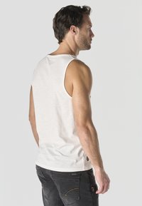 Hombre de cabello corto y oscuro, vestido con una camiseta blanca sin mangas y jeans oscuros, de pie y mirando hacia la derecha sobre un fondo liso.