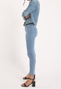Helle blaue Jeansjacke und enganliegende Jeans, kombiniert mit schwarzen Riemchensandalen mit Blockabsatz. Glatte Textur, maßgeschneiderte Passform, keine sichtbaren Muster.