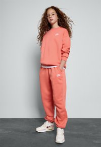 Koralen Nike sweatshirt in ujemajočih se trenirkah iz mehkega materiala, z belim logotipom na prsih in na levem hlačniku. Bele teniske z burgundskimi poudarki.