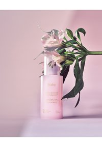 Huxley Skin Glow Essence in een zachte roze fles, met een zilveren pompdeksel, met bloemaccenten en afgedrukte productdetails. 50 mL.