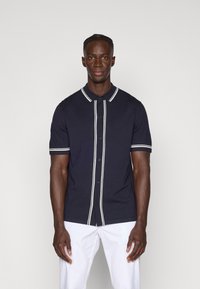 Polo de manga curta azul-marinho com detalhes em branco nos botões e riscas ao longo do colarinho e das mangas; feito de um tecido jersey suave.