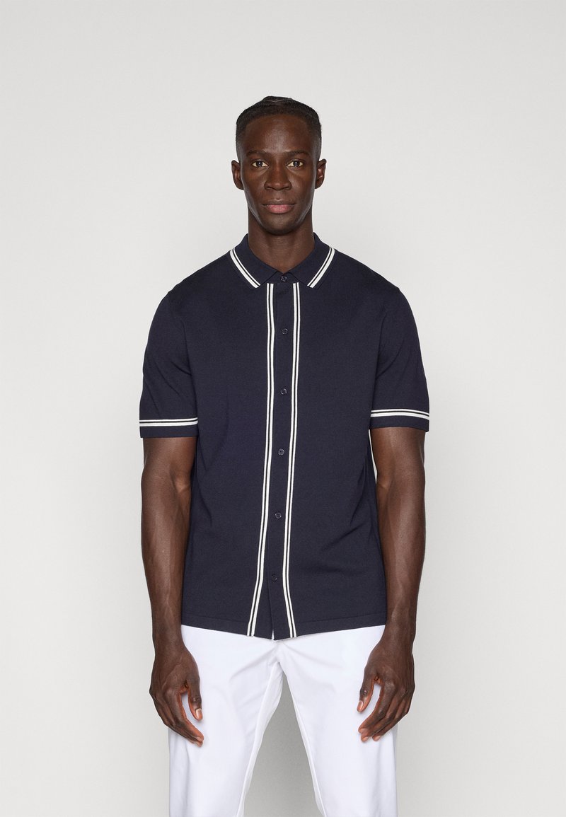 Polo de manga curta azul-marinho com detalhes em branco nos botões e riscas ao longo do colarinho e das mangas; feito de um tecido jersey suave.