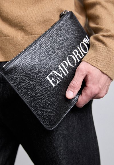 Mână ținând un pouch cu fermoar din piele texturată neagră Emporio, împotriva unui pulover bej și a unor pantaloni negri.