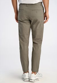 Pantalons chino kaki avec une coupe fuselée, dotés de deux poches arrière avec des boutons, fabriqués dans un tissu léger. Portés avec des baskets blanches.