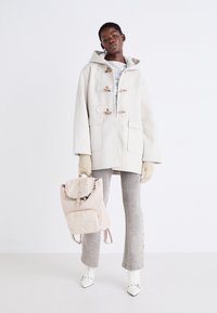 Manteau duffle à capuche gris clair avec des toggles en bois, associé à un pantalon gris côtelé et des talons blancs. Sac à dos beige avec des accents en fourrure.