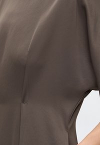 Gros plan d'une personne portant une chemise lisse marron avec des coutures visibles et une manche courte, sur un fond clair uni.