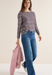 Pasiasty longsleeve w ciemnym fioletowym i jasnym lawendowym, w połączeniu z jasnoniebieskimi spodniami typu wide-leg. Różowa bluza trzymana jest w prawej ręce.