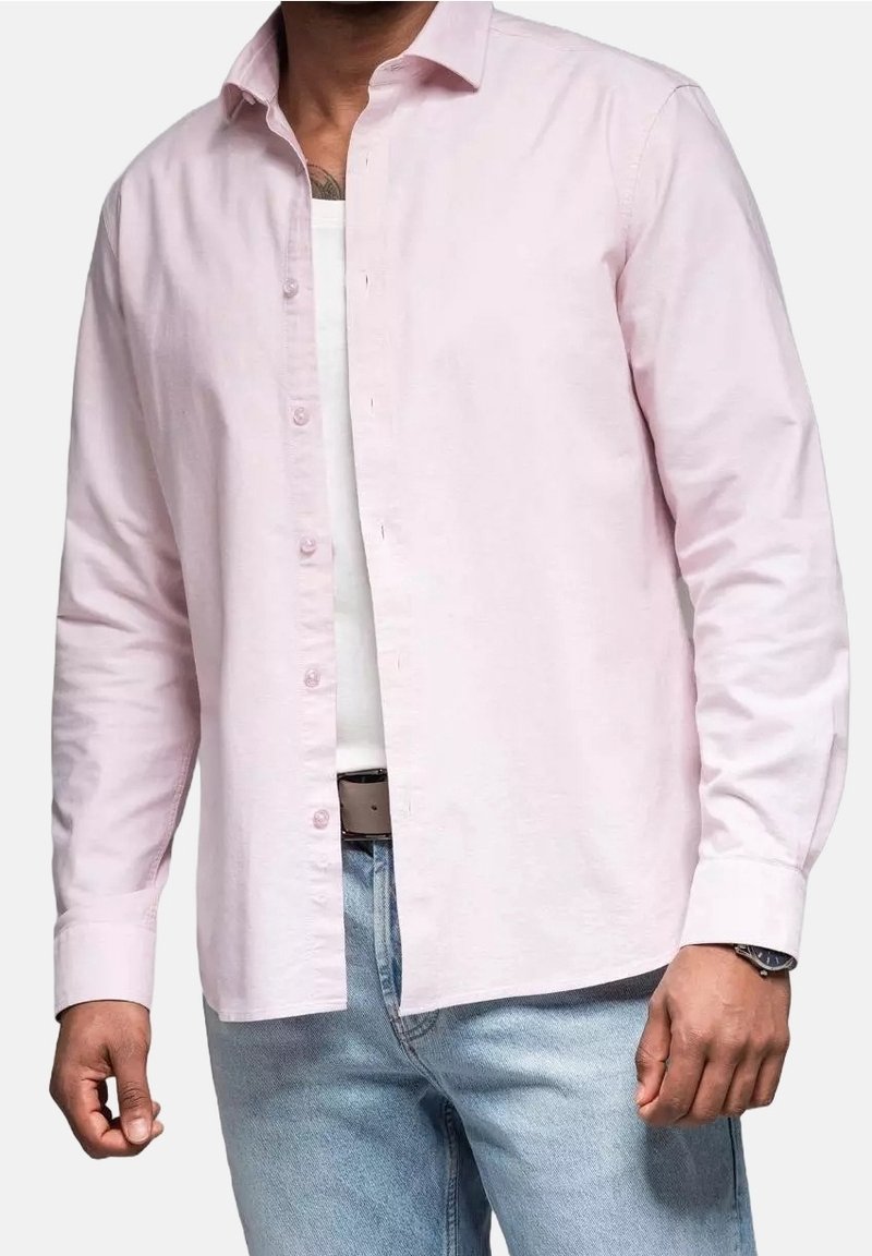 Homme portant une chemise rose clair à manches longues boutonnée sur un t-shirt blanc et un jean bleu clair, avec une montre au poignet gauche.
