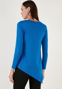 Top a maniche lunghe di un blu brillante, realizzato in tessuto morbido. Presenta un orlo asimmetrico e un colletto rotondo. Indossato con orecchini pendenti di grande effetto.