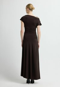 Ciemnobrązowa sukienka maxi wykonana z miękkiego materiału, z krótkimi rękawkami i marszczeniem w talii, sięgająca do podłogi, zestawiona z wysokimi obcasami.