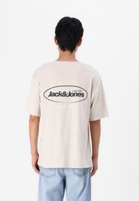 Beige bomull T-skjorte med en stor svart oval logo som leser "Jack & Jones Club1990" på baksiden, avslappet passform, korte ermer og rundede hem.