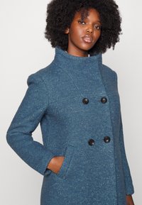 Manteau bleu à double boutonnage en tissu texturé, avec un col montant, des manches longues, des boutons noirs et des poches latérales.