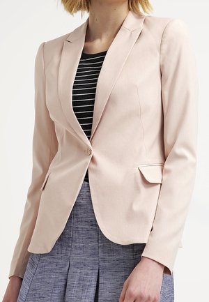 Kvinde iført en lyserød figursyet blazer over en sort og hvidstribet top, kombineret med et tekstureret gråt plisseret skørt.