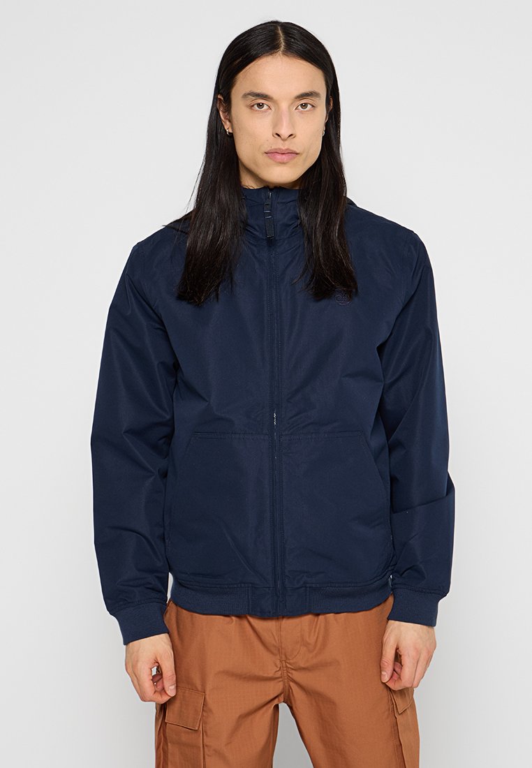 Mężczyzna z długimi czarni włosami, ubrany w zapinaną granatową kurtkę i brązowe spodnie cargo, stojący na tle jasnego, jednolitego tła.