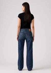 Tamsiai mėlyni džinsai su laisvu kirpimu, turintys šviesiai rudos odos etikėtę ant nugarinės juostos ir standartinius galinius kišenes. Dėvimas juodas trumpomis rankovėmis palaidinė.