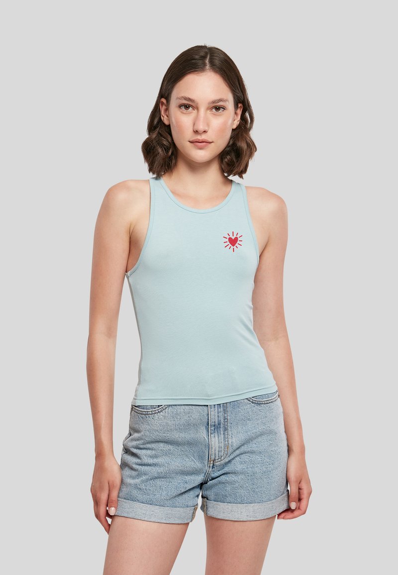 Jeune femme aux cheveux courts et bruns portant un débardeur bleu clair avec un cœur rouge imprimé et un short en jean retroussé, debout sur un fond gris.