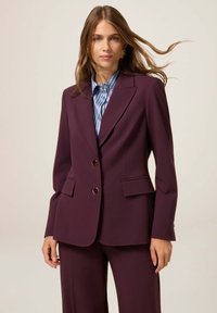 Donna con capelli lunghi, che indossa orecchini d'oro, un giubbotto viola con bottoni, pantaloni coordinati e una camicia a righe blu su uno sfondo uniforme.