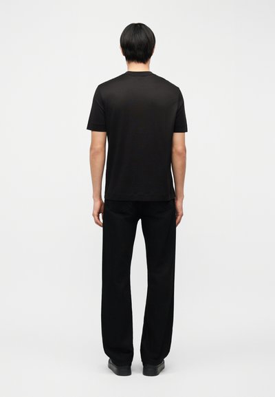 Άντρας που στέκεται με την πλάτη γυρισμένη, φοράει μαύρο απλό κοντομάνικο μπλουζάκι, μαύρο παντελόνι και μαύρα παπούτσια σε λευκό φόντο.