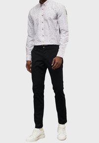 Camicia a bottoni con motivo chiaro e bottoni marroni, abbinata a pantaloni neri slim-fit e sneakers bianche. Texture morbide evidenti nel tessuto.