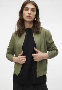 Maslinasto zeleni bomber jakna s zlatnim patent-zatvaračem, rebrastim manžetama i mekanom teksturom, nosi se preko crnog pulovera. Model pozira s rukama na patent-zatvaraču.
