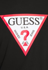 Czarna koszulka z dużym logo Guess w czerwono-białym trójkącie. Projekt zawiera tekst "WASHED", znak zapytania i numeryczne akcenty.