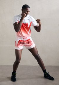 Boxer in red and white sportswear and black shoes stands in fighting stance with fists up against a plain wall background.